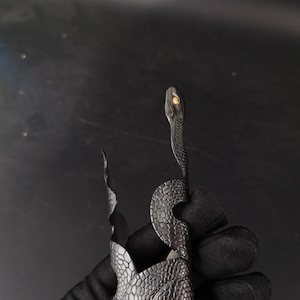 May include: A black metal snake-shaped bracelet with a textured, scale-like pattern. The snake's head is raised, and its eye is a golden colour. The bracelet has a unique, artistic design.