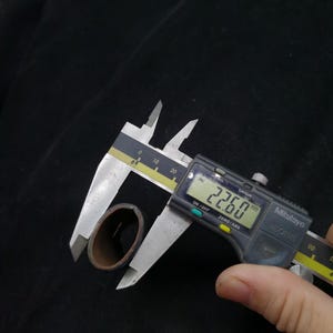 May include: A small, dark, cylindrical metal tube being measured by a digital Mitutoyo calliper. The display on the silver and black measuring tool clearly shows a reading of 22.60 mm, indicating the precise outer diameter of the circular object.