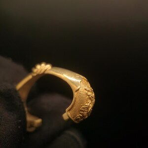 May include: A gold-colored open cuff bracelet. The bracelet features detailed floral and textured patterns on the outer surface. The open end design allows for easy wear. The bracelet is shown against a dark background.