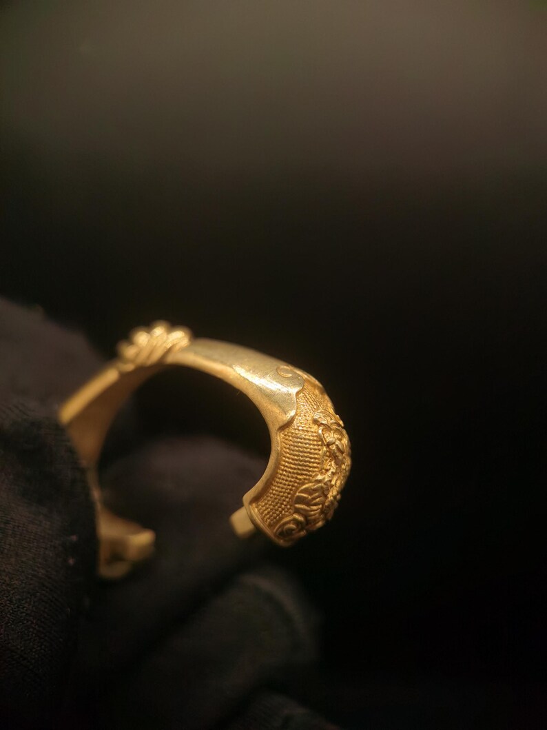 May include: A gold-colored open cuff bracelet. The bracelet features detailed floral and textured patterns on the outer surface. The open end design allows for easy wear. The bracelet is shown against a dark background.