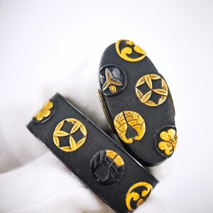 May include: Two black metal sword fittings featuring a textured surface and raised gold-coloured circular emblems. The oval-shaped piece and the rectangular band display various floral and geometric motifs in relief against the dark background.