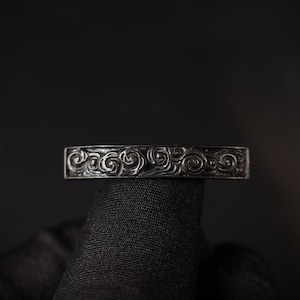 Könnte beinhalten: Ein silberner Ring mit einem detaillierten, wirbelnden Muster. Der Ring wird vor einem dunklen Hintergrund präsentiert, der das komplizierte Design hervorhebt. Das Band weist eine durchgehende Reihe stilisierter floraler oder wellenartiger Motive auf.