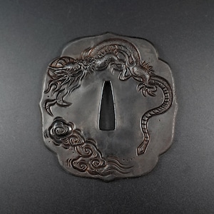May include: An antique Japanese tsuba, or sword guard, with a detailed dragon design. The dragon is intricately carved in relief, with a cloud motif. The tsuba is made of dark metal, probably iron or steel, and has a central opening.