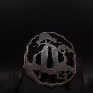 May include: A dark, circular metal tsuba featuring an intricate openwork design of a pine tree. The sword guard has a textured, matte finish with a central elongated opening for the blade and a smaller circular opening for a utility tool.