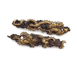 May include: Two ornate, antique-style dragon sculptures. The dragons are gold-colored with intricate details, set against a dark, possibly oxidized, background. The dragons are in a dynamic pose, suggesting movement.