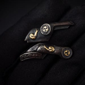May include: Two silver-toned metal accessories with gold detailing. Each piece features a snail and a circular design. The accessories are set against a dark background, highlighting their intricate details and craftsmanship.