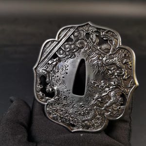 May include: An intricately carved silver-coloured Japanese tsuba, a sword guard, depicting a dragon in relief amongst swirling waves and clouds. The tsuba has a central opening for the sword tang and is held by a gloved hand.