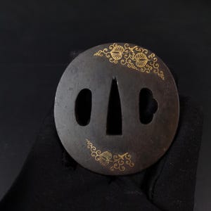 May include: A circular dark metal tsuba featuring gold-coloured inlaid leaf patterns. The sword guard has a central tapered opening for the blade and two smaller side apertures, one oval and one shaped like a stylised clover or petal.