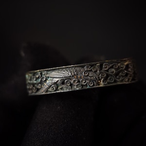 May include: A dark silver-toned metal ring featuring an intricate, raised relief design of a bird surrounded by swirling, vine-like patterns. The metal has a weathered, oxidized finish with visible dark patina in the recessed areas of the carving.
