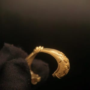May include: A gold-colored bracelet with intricate embossed patterns. The open-ended design features detailed floral and geometric motifs, suggesting a vintage or antique style. The bracelet is held against a dark background.