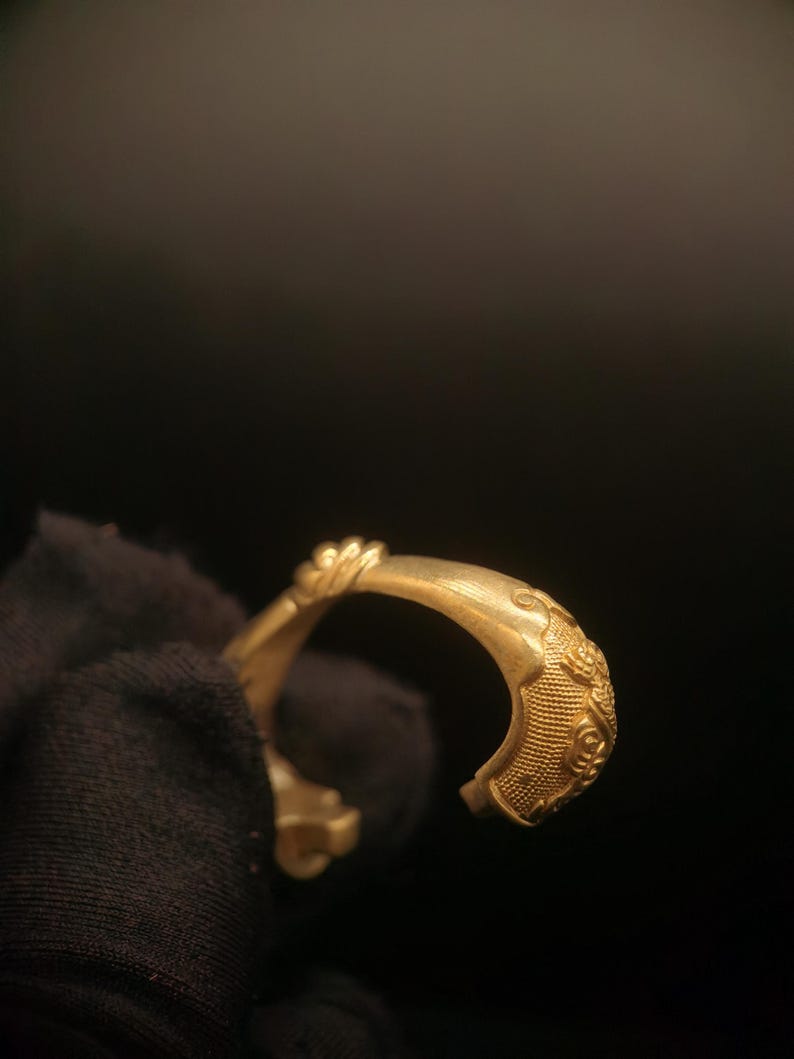 May include: A gold-colored bracelet with intricate embossed patterns. The open-ended design features detailed floral and geometric motifs, suggesting a vintage or antique style. The bracelet is held against a dark background.