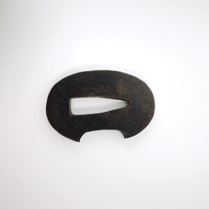 Peut inclure: Une tsuba en métal sombre et patiné de forme ovale, présentant une découpe rectangulaire centrale effilée et une petite encoche semi-circulaire sur le bord inférieur. La surface de la garde d'épée montre une texture vieillie et oxydée.