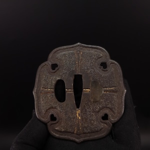 May include: A dark, metallic tsuba sword guard with a lobed, quatrefoil shape. The surface features intricate, fine-line floral engravings and gold-coloured inlay accents. It includes a central tapered opening and two smaller side apertures.