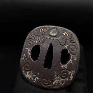 May include: A dark metal sword guard, or tsuba, featuring an irregular rounded shape with a central triangular opening and two smaller side apertures. The surface is decorated with embossed cloud motifs accented with gold and silver metallic inlays.