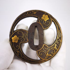 Peut inclure: Une garde d'épée circulaire en métal sombre, appelée tsuba, dotée d'une ouverture centrale ovale. La surface est ornée de motifs floraux et de volutes dorés incrustés, contrastant avec la base en métal sombre et texturé.