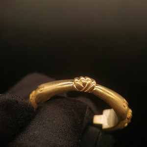 May include: A gold-colored bracelet with a decorative knot design at the center. The bracelet has a curved shape and appears to be made of metal. The design includes small, raised details near the ends. The background is dark.