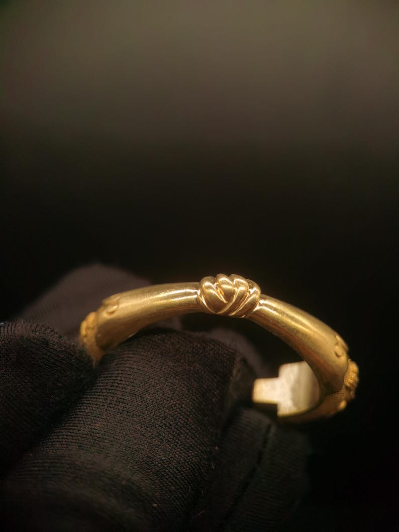 May include: A gold-colored bracelet with a decorative knot design at the center. The bracelet has a curved shape and appears to be made of metal. The design includes small, raised details near the ends. The background is dark.