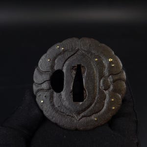 May include: A dark brown, circular metal tsuba featuring a scalloped edge and a textured surface. The piece includes a central tapered opening, a smaller circular side hole, and several small, inlaid gold-coloured dots scattered across the face.