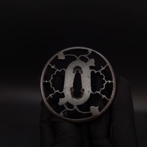 May include: A circular dark metal tsuba featuring an openwork design of stylised leaves and vines surrounding a central elongated oval opening. The metal surface has a matte, weathered texture with visible signs of age and oxidation throughout.