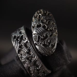 May include: A silver ring featuring a detailed wave pattern. The ring comprises an oval-shaped top and a band, both intricately carved. The design is set against a dark backdrop, accentuating the metallic finish. The ring's design evokes a sense of the ocean.