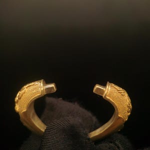 May include: A gold-colored, open-ended bracelet with detailed, textured designs on the curved ends. The bracelet has a polished finish and is set against a dark background, highlighting its metallic sheen. The design appears to be inspired by a classical or vintage aesthetic.