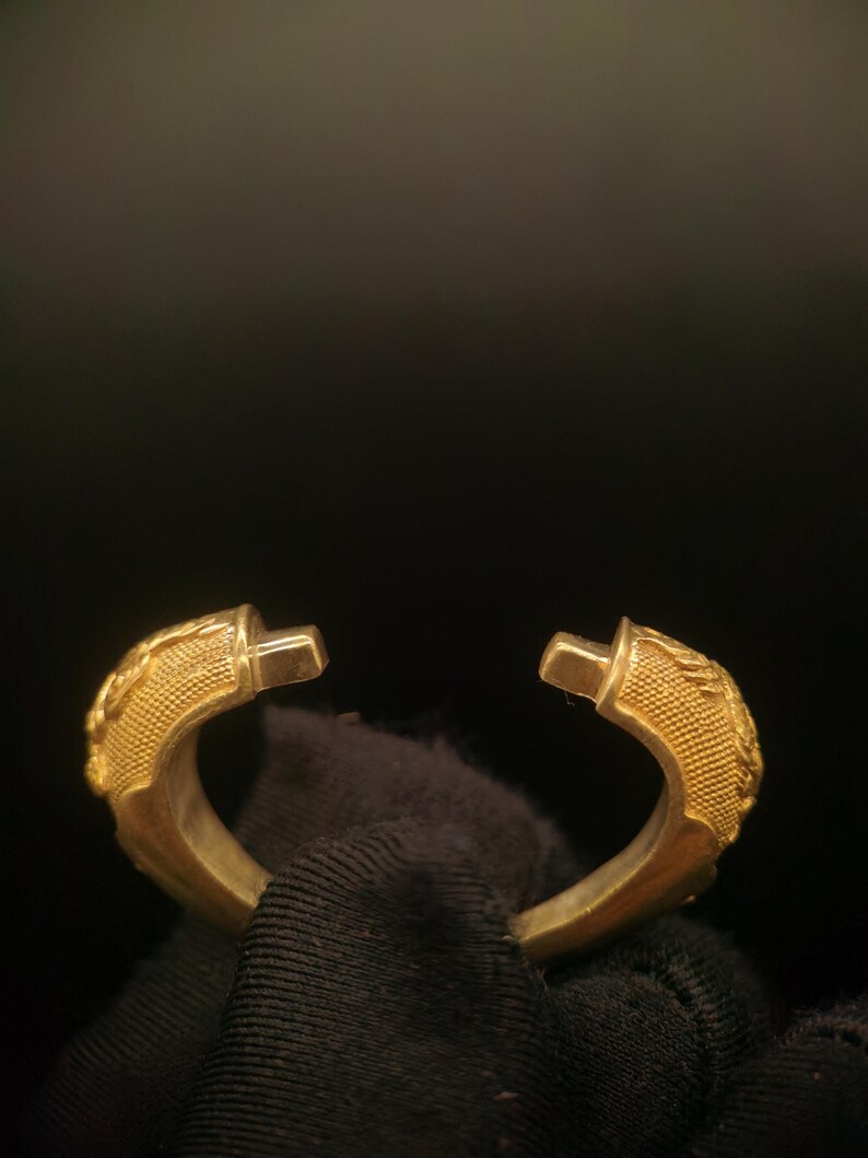 May include: A gold-colored, open-ended bracelet with detailed, textured designs on the curved ends. The bracelet has a polished finish and is set against a dark background, highlighting its metallic sheen. The design appears to be inspired by a classical or vintage aesthetic.