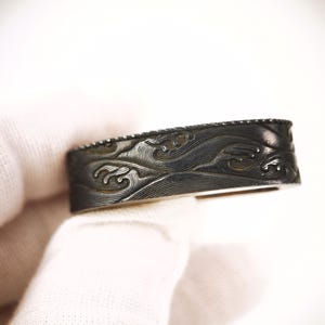 May include: A dark, rectangular metal object featuring intricate, engraved wave patterns across its surface. The piece has a smooth, polished finish with deep grooves that highlight the flowing, rhythmic lines of the carved design.