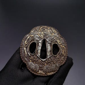 May include: A circular metal tsuba featuring a dark, weathered patina with raised relief carvings of a phoenix and foliage. The sword guard has a central elongated opening flanked by two smaller oval apertures, all with silver-toned accents.