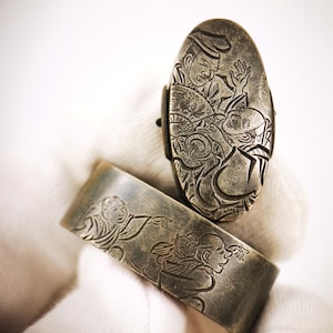 May include: An oval-shaped metal object and a curved metal band, both featuring detailed engraved illustrations of figures in traditional attire. The metal has a dark, aged patina finish with deep grooves highlighting the intricate character designs.