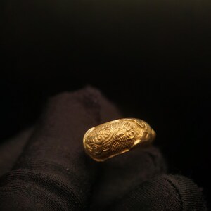 May include: A gold ring with an ornate, textured design. The ring features a repeating pattern of swirls and lines, creating a detailed and eye-catching appearance. The ring is held against a dark background.