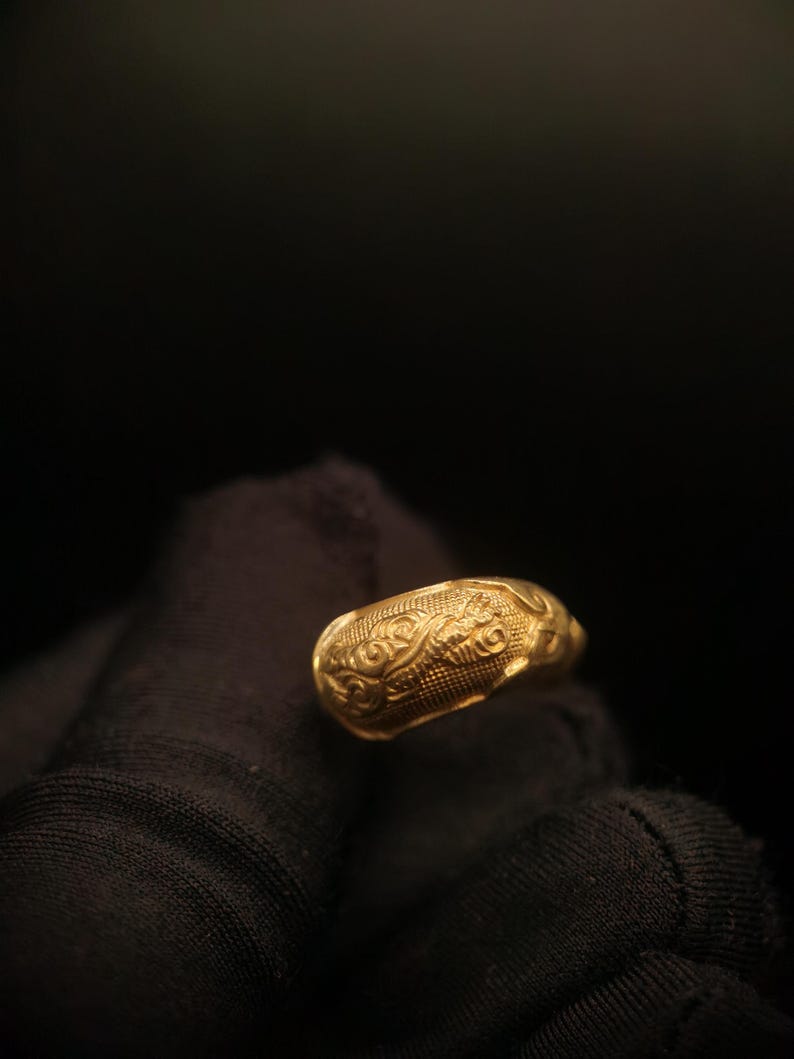 May include: A gold ring with an ornate, textured design. The ring features a repeating pattern of swirls and lines, creating a detailed and eye-catching appearance. The ring is held against a dark background.