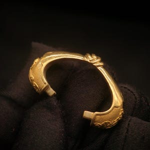 May include: A gold-colored cuff bracelet with ornate detailing. The bracelet features a floral design and textured patterns. The open design allows for easy wear. The bracelet is shown against a dark background.
