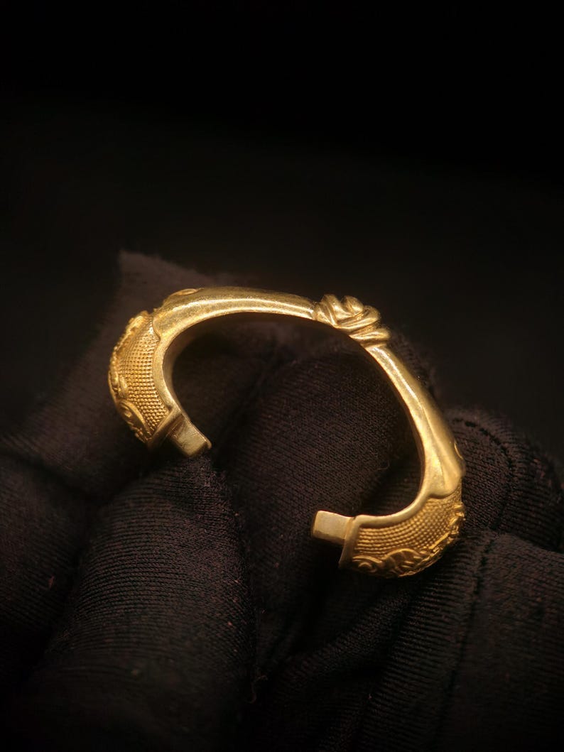 May include: A gold-colored cuff bracelet with ornate detailing. The bracelet features a floral design and textured patterns. The open design allows for easy wear. The bracelet is shown against a dark background.