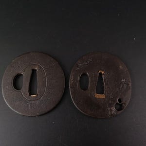 Peut inclure: Deux gardes de sabre tsuba circulaires en métal brun foncé. Chaque pièce présente une ouverture rectangulaire centrale effilée et un petit trou circulaire adjacent. Les surfaces ont une finition texturée et patinée par le temps.