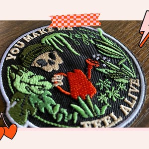 Könnte beinhalten: Gestickter Patch mit einem Schädel, der eine Mütze trägt und eine Gießkanne hält. Der Schädel ist von grünen Pflanzen umgeben und der Text "YOU MAKE ME FEEL ALIVE".