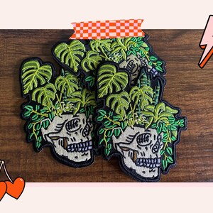 Könnte beinhalten: Gestickte Aufnäher mit einem Totenkopf, aus dem grüne Pflanzenblätter sprießen. Die Aufnäher haben einen schwarzen Rand und sind mit grünem und beigem Garn verziert. Die Aufnäher werden auf einer Holzoberfläche präsentiert.