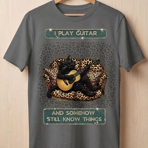 May include: Gray t-shirt featuring a black cat playing a guitar on a leopard-print couch. The text "I PLAY GUITAR" and "AND SOMEHOW STILL KNOW THINGS" are displayed above and below the cat illustration.