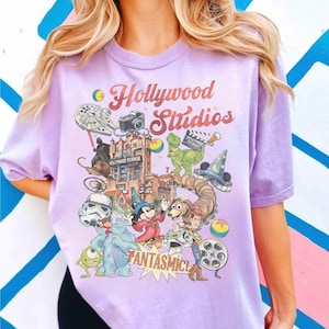 May include: A light purple t-shirt with a colorful graphic featuring the words "Hollywood Studios" and various cartoon characters and icons. The design includes a Star Wars ship, a camera, and a dinosaur.