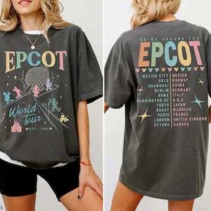 Może przedstawiać: Ciemnoszara koszulka EPCOT World Tour z kolorową grafiką z przodu i z tyłu. Z przodu znajduje się logo EPCOT, globus i postacie z kreskówek. Z tyłu lista miast na całym świecie. Zał. 1982.