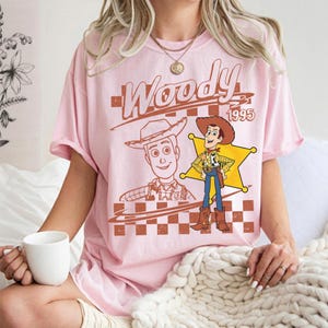 May include: A light pink t-shirt featuring a graphic of the character Woody from the movie Toy Story. The design includes the word "Woody" in a brown font, the year "1995", and two illustrations of the character. The shirt has short sleeves.