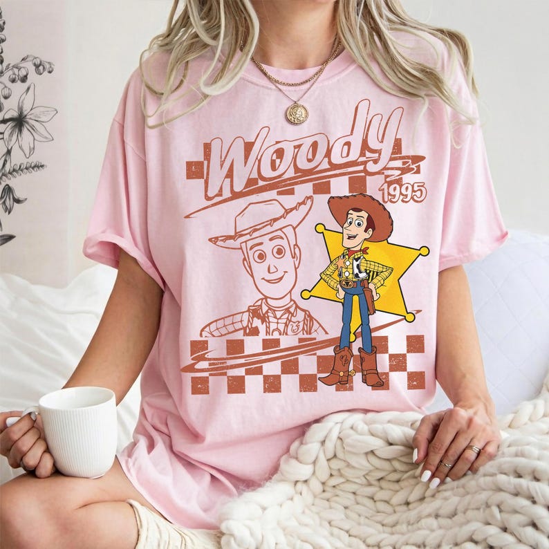 May include: A light pink t-shirt featuring a graphic of the character Woody from the movie Toy Story. The design includes the word "Woody" in a brown font, the year "1995", and two illustrations of the character. The shirt has short sleeves.