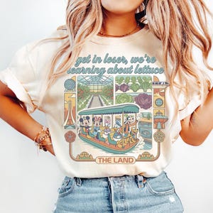 Könnte beinhalten: Cremefarbenes T-Shirt mit einem Grafikdesign im Retro-Stil. Der Aufdruck zeigt den Text "get in loser, we're learning about lettuce", ein Boot mit Disney-Figuren, ein Gewächshaus und Gemüse. Ein lässiges Kleidungsstück.