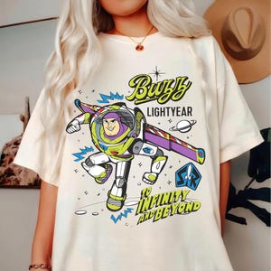 Puede incluir: Camiseta color crema con un diseño gráfico de Buzz Lightyear en una pose dinámica. El diseño incluye las palabras "Buzz Lightyear" y "To Infinity and Beyond" en una fuente divertida, con elementos espaciales.