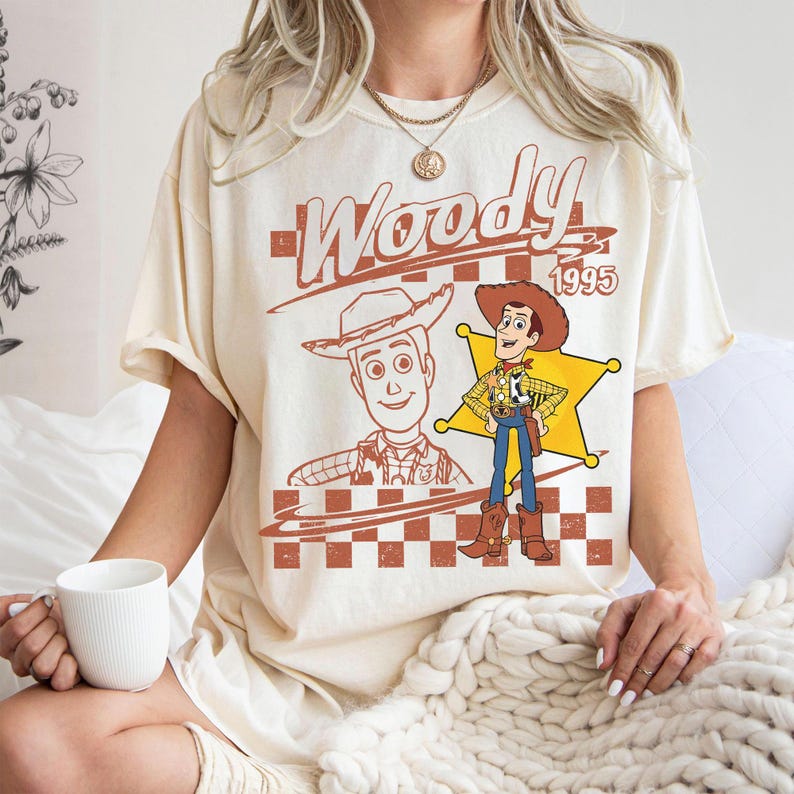 May include: Cream-colored t-shirt featuring a graphic of the character Woody from the movie Toy Story. The design includes the word "Woody" in a brown, stylized font, the year "1995", and two illustrations of the character.