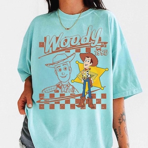 May include: A light blue t-shirt featuring a graphic of the character Woody from the movie Toy Story. The design includes the word "Woody" in a brown font, the year "1995", and a sheriff's badge.