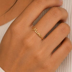 May include: A gold-toned ring featuring the word Mama written in a delicate, cursive script font. The ring has a thin, polished band that encircles the finger, showcasing a minimalist design with the lettering integrated directly into the band.