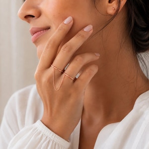 Peut inclure: Une double bague en or rose reliée par une fine chaîne délicate. Les deux anneaux circulaires lisses sont portés sur des doigts adjacents, avec la chaîne fine qui se drape entre eux pour créer un aspect lié et superposé sur la main.