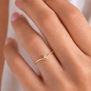 May include: A thin gold ring featuring a V-shaped band and a single small, round, clear gemstone set at the centre point. The metal has a smooth, polished finish, and the design follows a simple, pointed chevron silhouette on the finger.