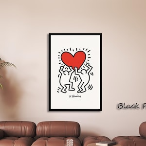 May include: A framed wall art print featuring a black and white line drawing of two figures holding a large red heart above their heads. The artwork includes the signature K. Haring at the bottom and is enclosed in a simple black frame.