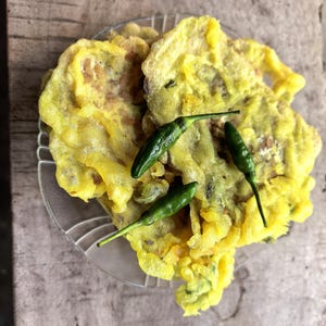 May include: A stack of golden-yellow, deep-fried vegetable fritters on a clear glass plate, topped with three small, glossy, dark green bird's eye chili peppers. The fritters have a crispy, textured batter coating with visible green herbs.